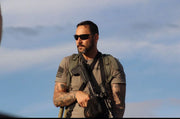 Tätowierter Mann mit Sonnenbrille, taktischer Ausrüstung und Gewehr vor blauem Himmel
